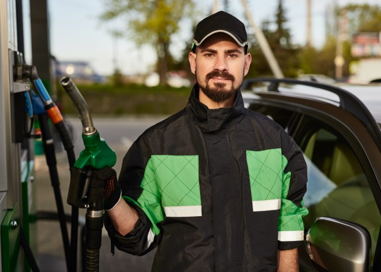 Confident bearded male worker at gas station in cap standing near car and looking at camera