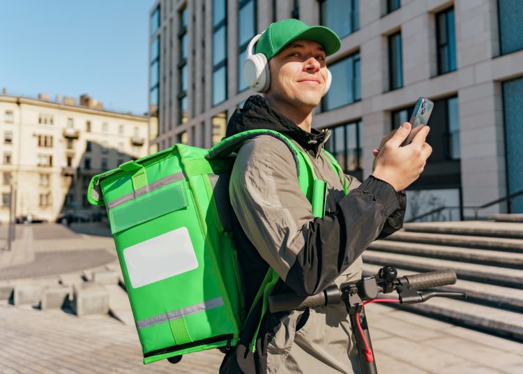 Smiling delivery man with green backpack checking smartphone in urban setting, headphones on.